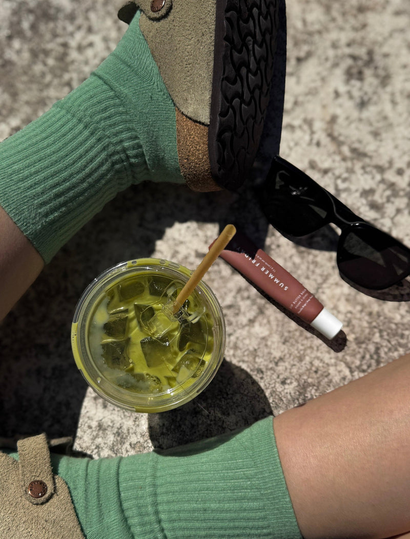 Person holding a drink with a straw, wearing green socks and brown shoes, on a textured surface.