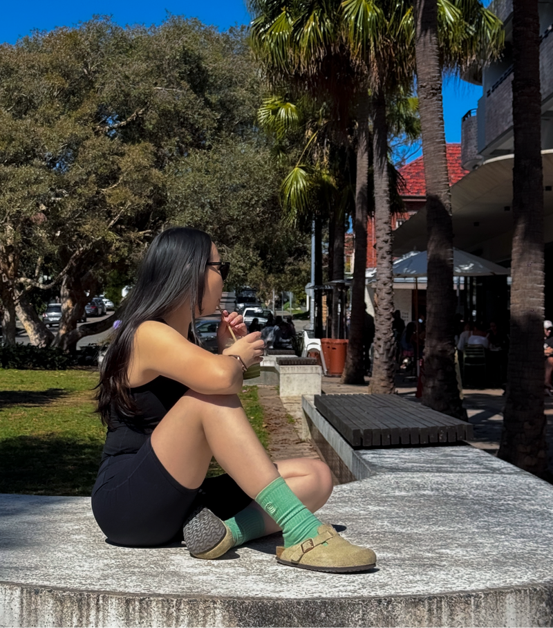 Woman sitting on a stone bench in a park with palm trees and buildings in the background.
