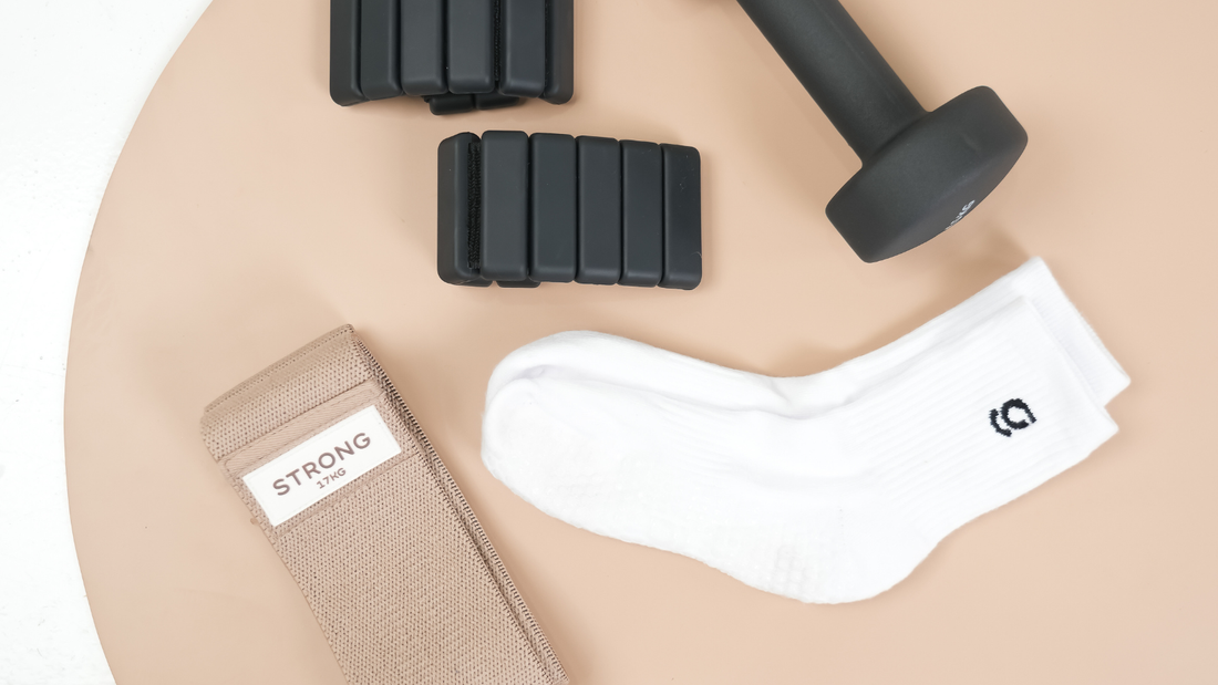 Gravi grip socks, resistance band, ankle weights and a black dumbbell on a beige mat