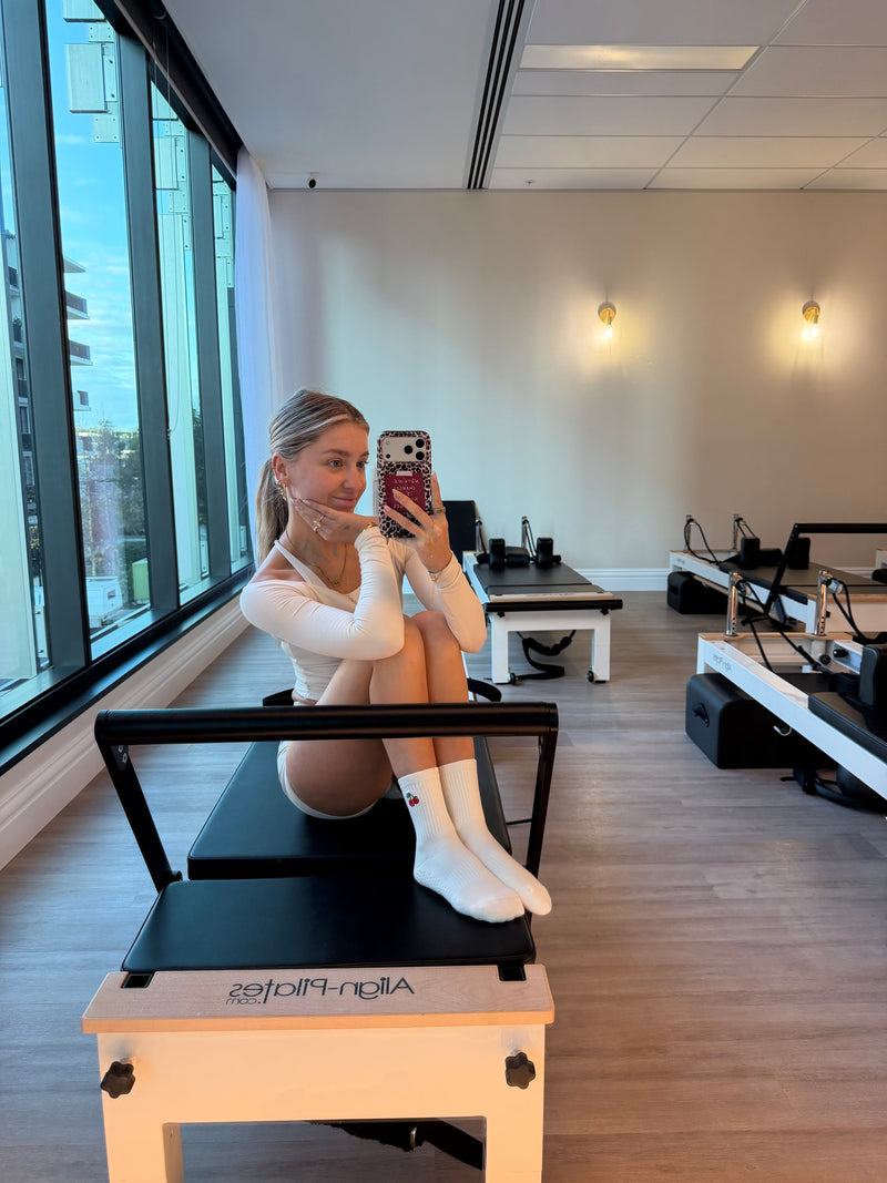 A woman takes a mirror selfie in a Pilates studio while seated on a reformer machine. The room has light wood floors, beige walls, large windows on the left, and other Pilates equipment in the background. Wearing the Cherry Crew Grip Socks.
