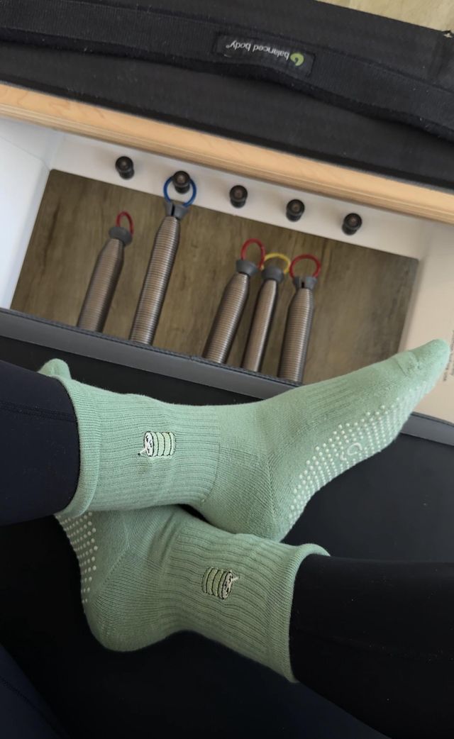 A pair of gravi matcha crew rip socks worn on feet, shown in a Pilates studio setting with a reformer machine and resistance tubes in the background. 