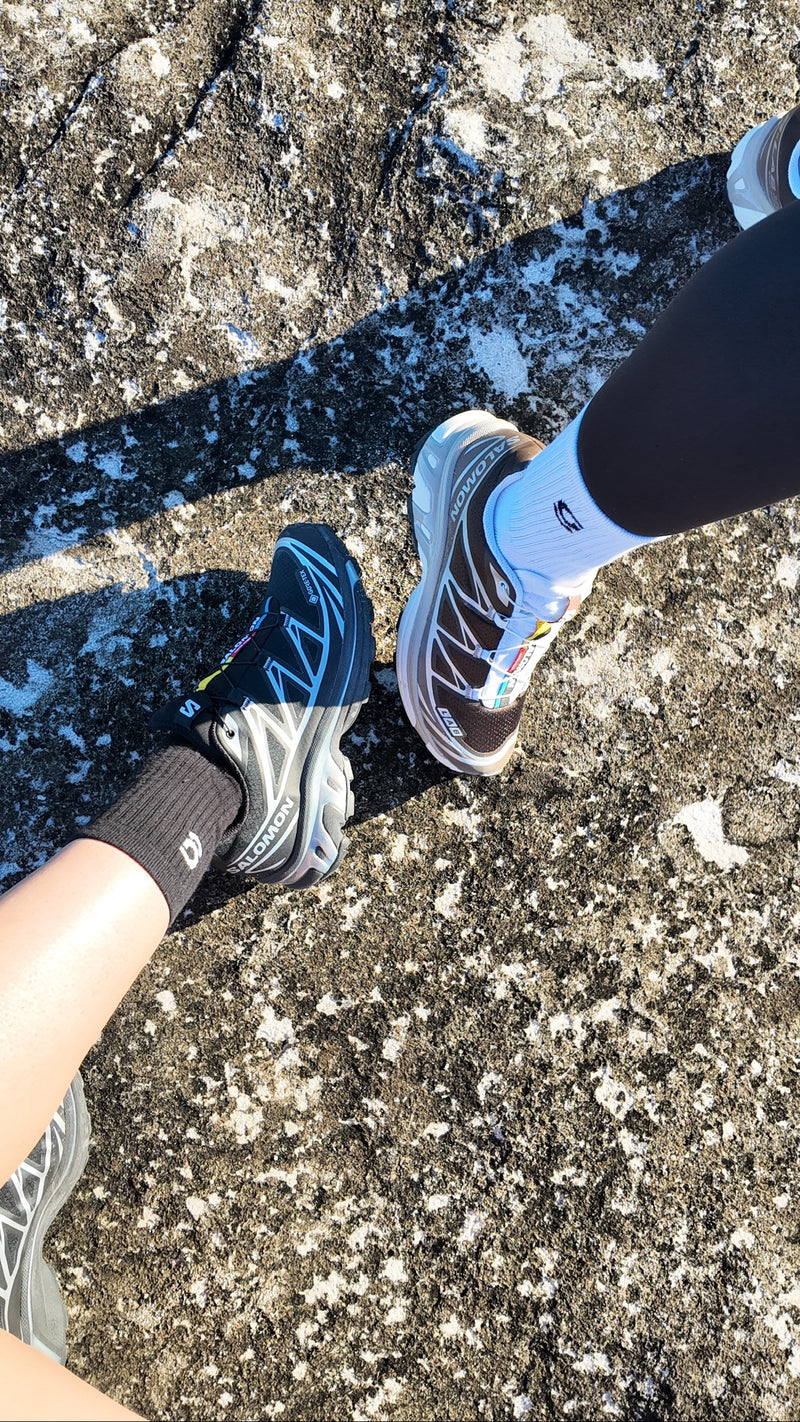 A top-down view of two people's legs and feet standing on a rocky, snowy outdoor surface. One person wears a black Salomon trail running shoe with gravi black grip socks, and the other wears a brown and white trail running shoe with white gravi grip socks.