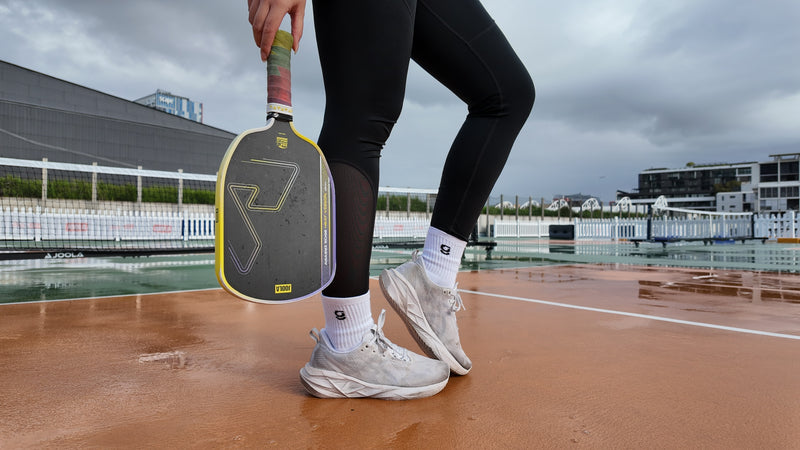 A person standing on a wet pickleball court holding a paddle, wearing black leggings, white gravi grip socks, and white sneakers. The background shows an outdoor pickleball court with buildings and overcast sky.